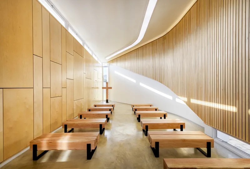 Chapel interior with timber benches on polished concrete floor beneath curved ceiling with continuous light slot
