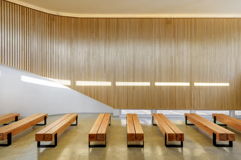 Vertical timber slat wall with five horizontal light strips above rows of wooden pews on concrete floor