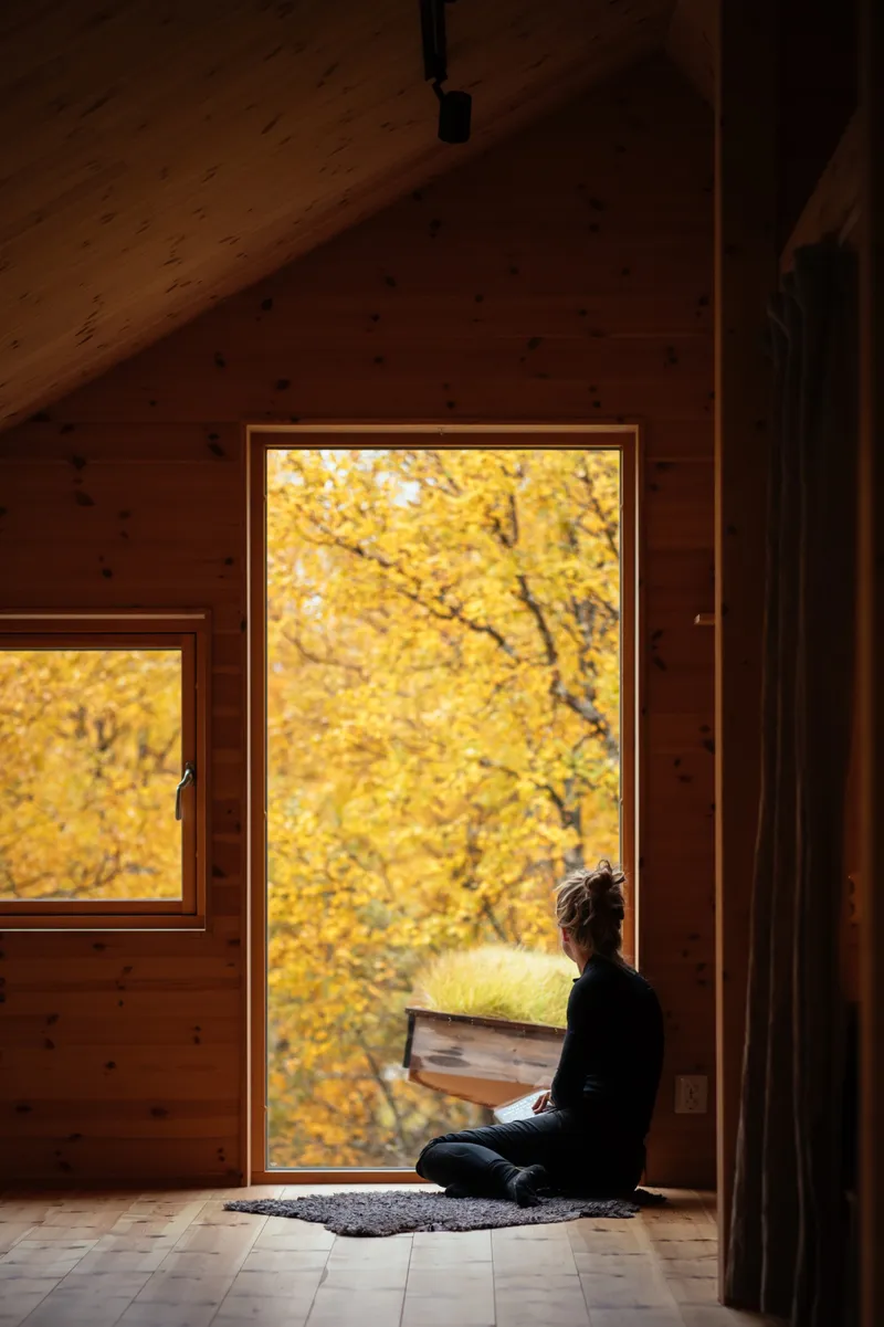 Person sitting in a wood-clad window alcove overlooking yellow autumn foliage outside