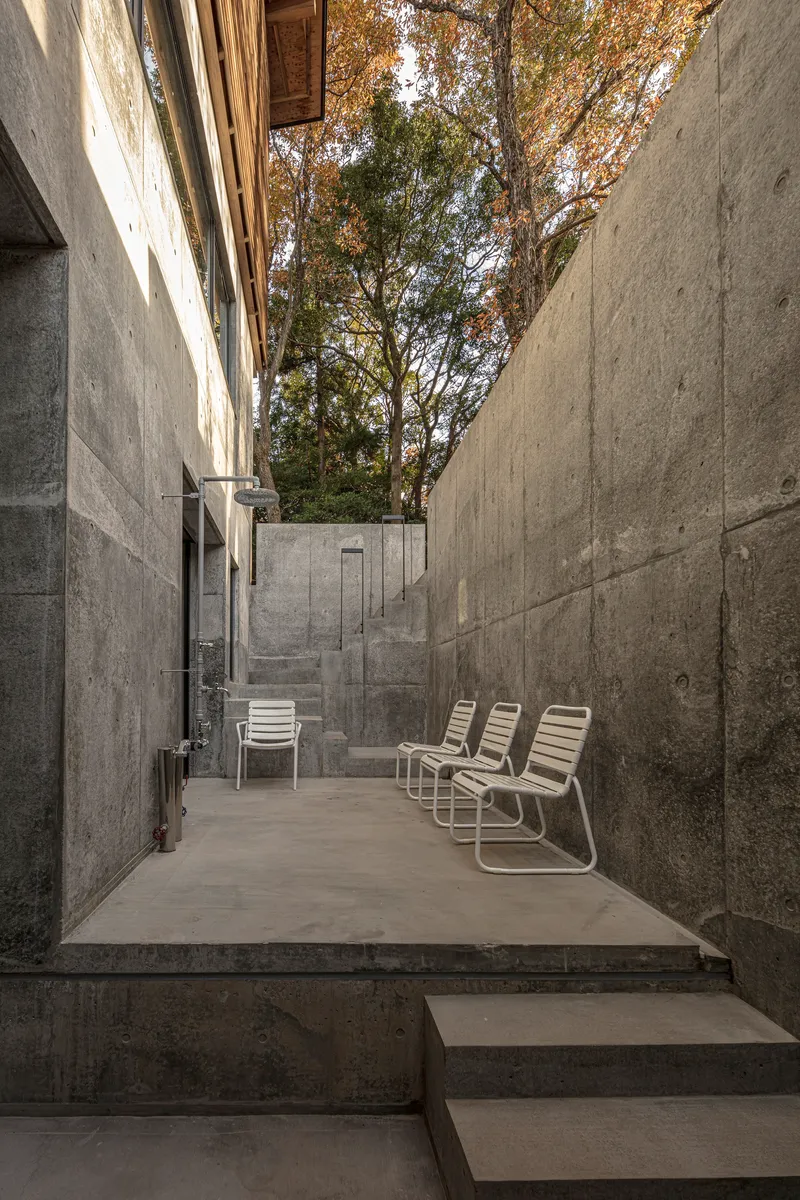 Concrete courtyard with metal lounge chairs flanked by board-formed walls beneath autumn trees