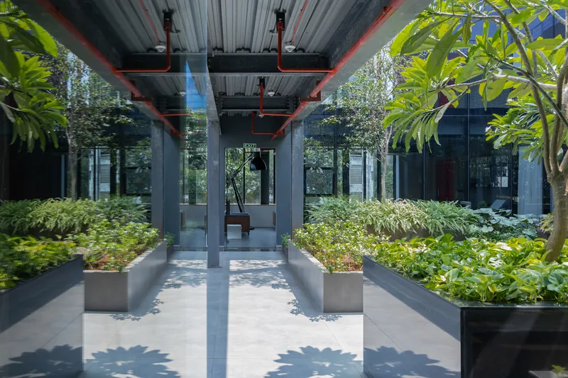 Covered walkway with exposed corrugated ceiling and red structural supports framing planted beds and trees in dappled sunlight