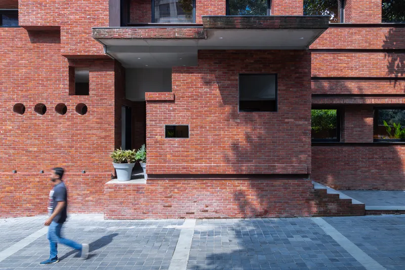 Red brick facade with protruding volumes and punched windows as a person walks across the paved forecourt