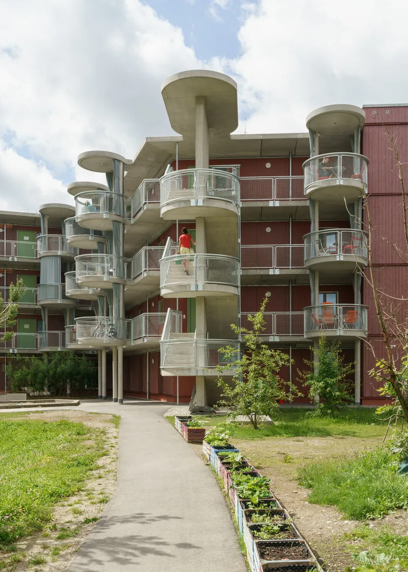 Concrete balcony columns with mushroom capitals rising from a courtyard garden with young trees and planting beds