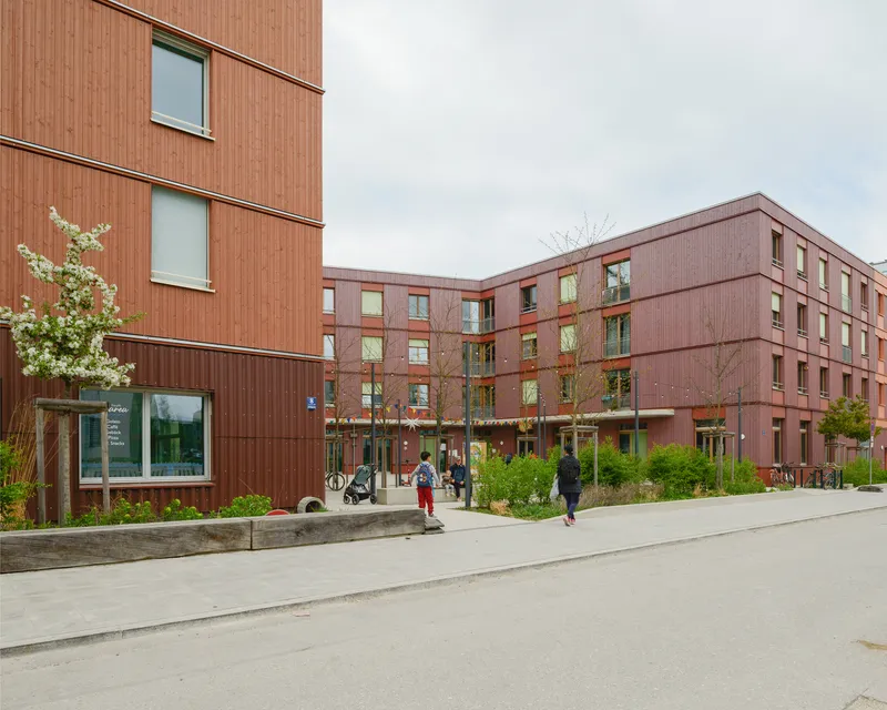 Red corrugated metal facade with residents walking along a paved courtyard under an overcast sky