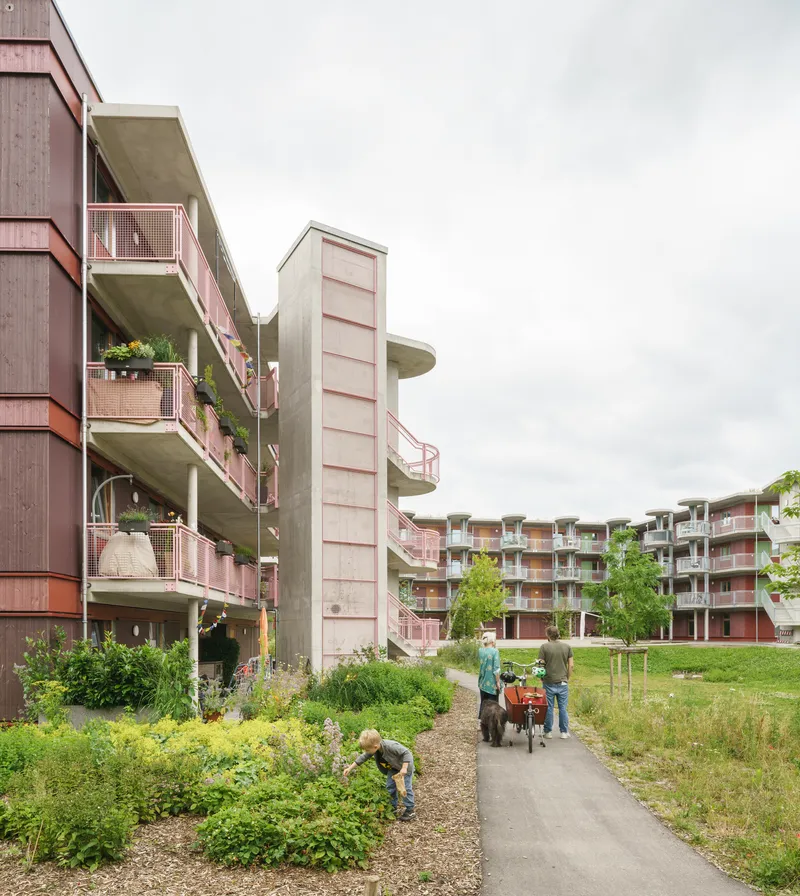 Curved residential blocks with exposed concrete access balconies flanking a gravel pathway through community gardens with residents tending plants