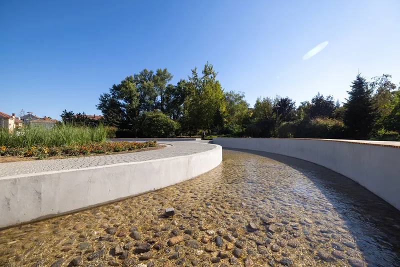 Curved concrete retaining wall with planted bed enclosing a shallow water channel with pebbled bottom