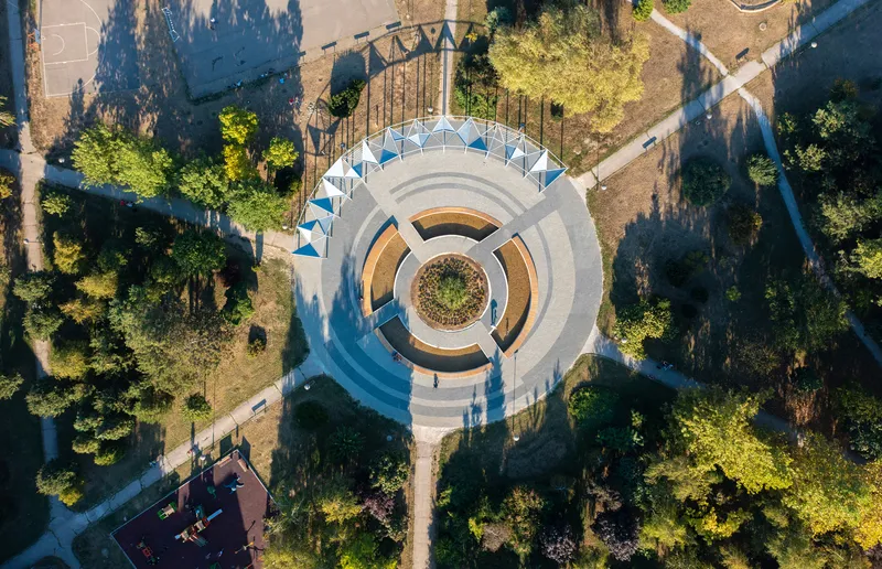 Drone view of the circular pavilion with blue canopy panels nestled among trees casting long shadows