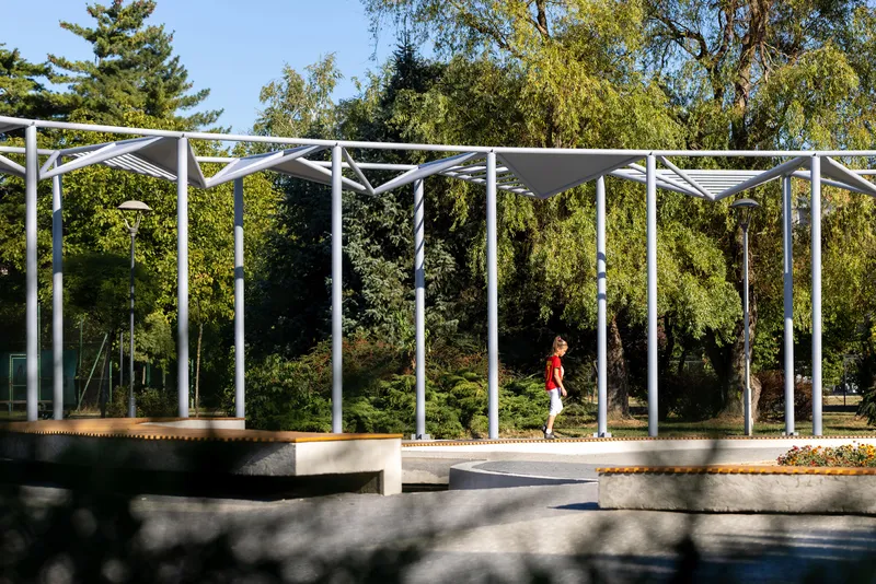 Person walking along the raised concrete platform under the steel pergola with trees in background