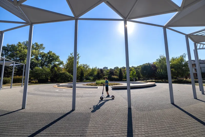 View through white steel columns and triangular canopy panels toward a paved plaza with a person on a scooter