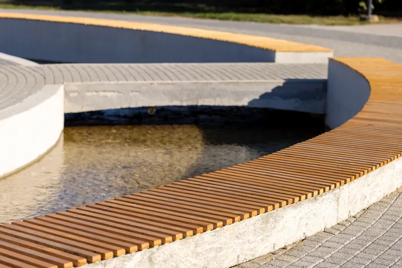 Slatted timber bench curving along the edge of a shallow concrete water feature in afternoon sun
