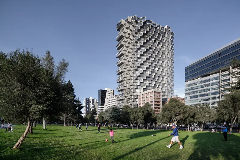Pixelated concrete balcony facade rising above a park lawn with children playing under clear skies