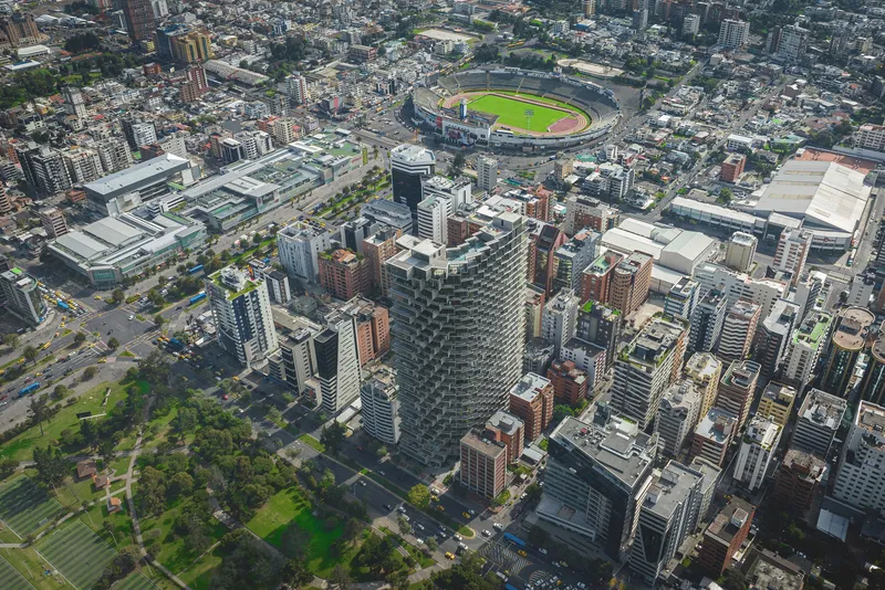 Drone view showing the tower rising above dense urban blocks with a stadium in the background