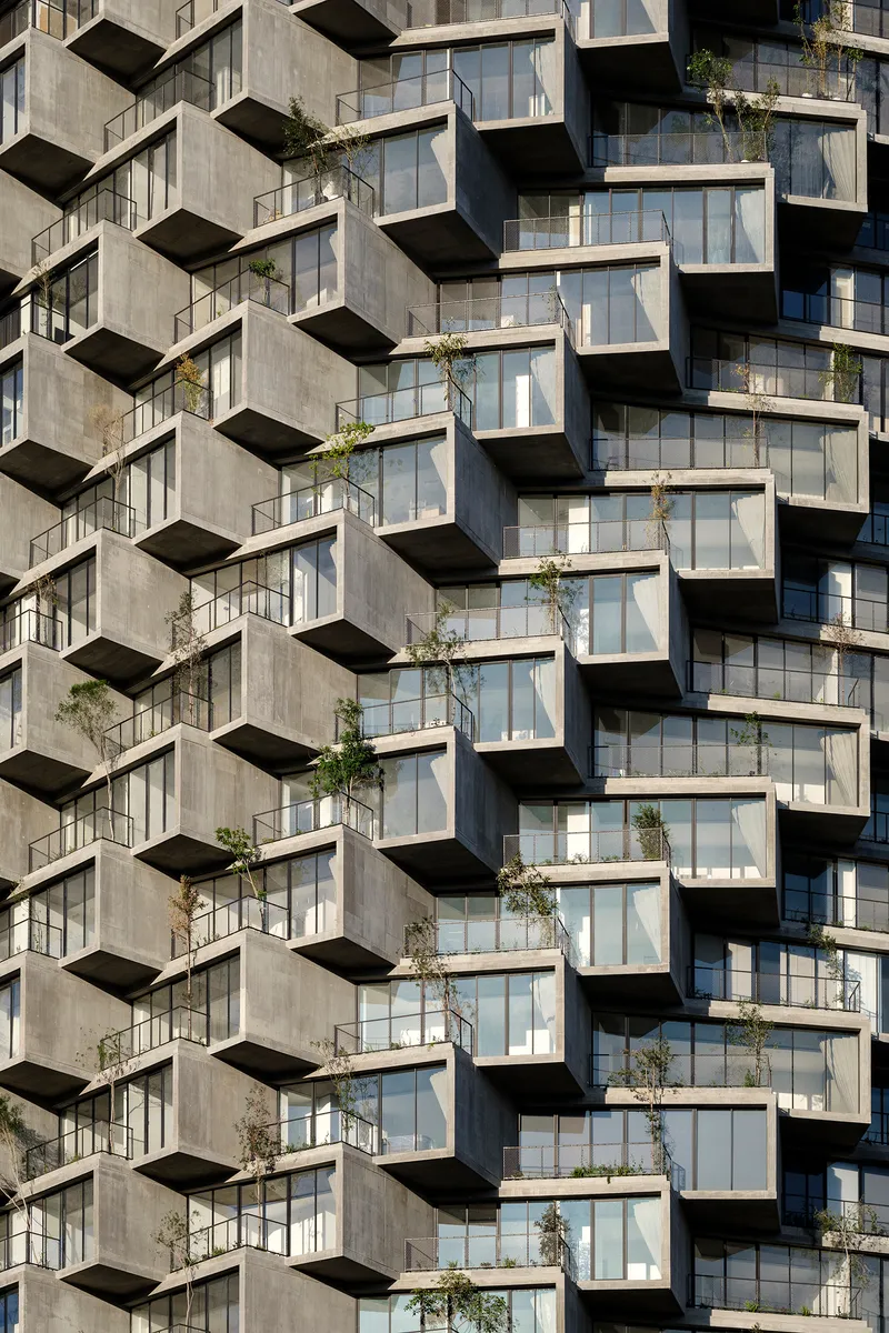 Concrete facade with staggered balconies and floor-to-ceiling glazing planted with young trees in bright sunlight