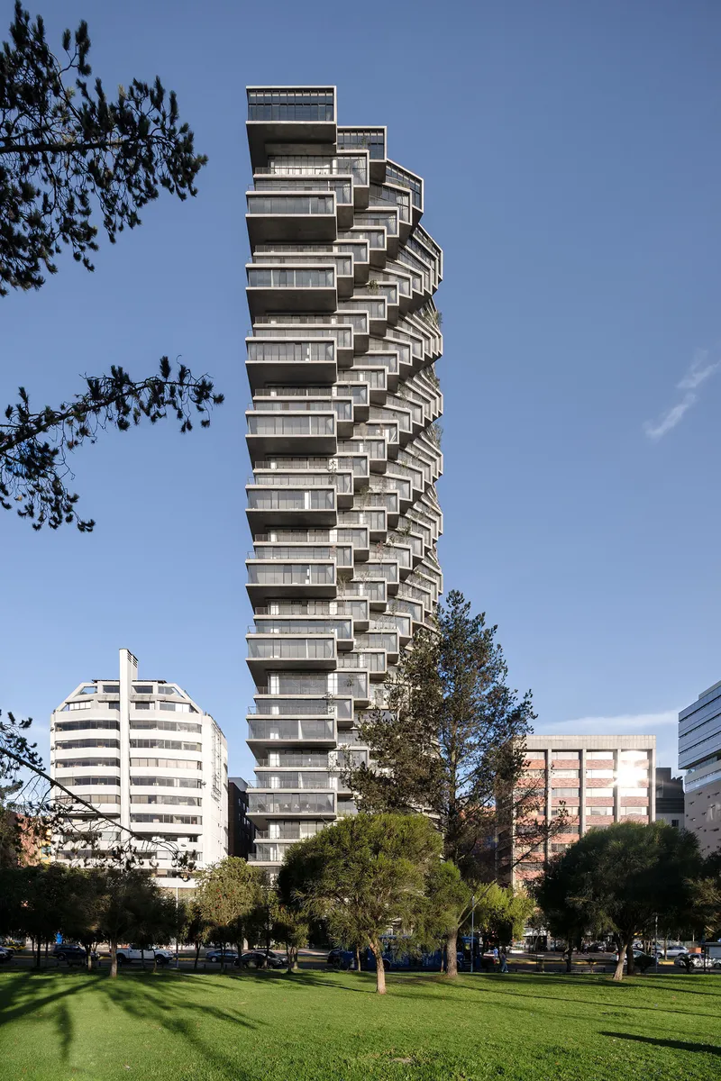 Twisting residential tower with offset balconies rising above a landscaped park under clear blue sky