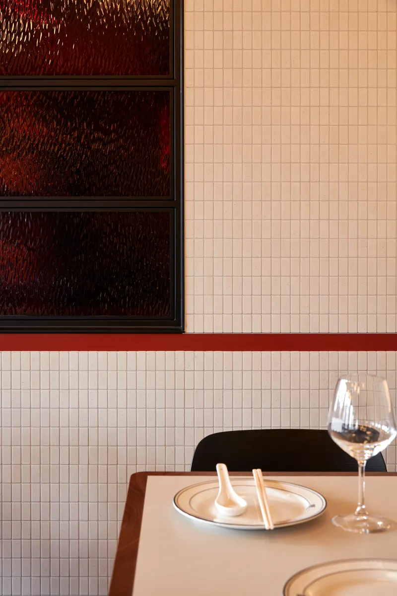 Detail of white mosaic tile wall with red trim meeting dark textured glass display shelves