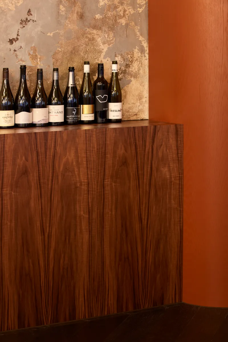 Walnut veneer cabinet with wine bottles displayed against weathered plaster wall in natural light