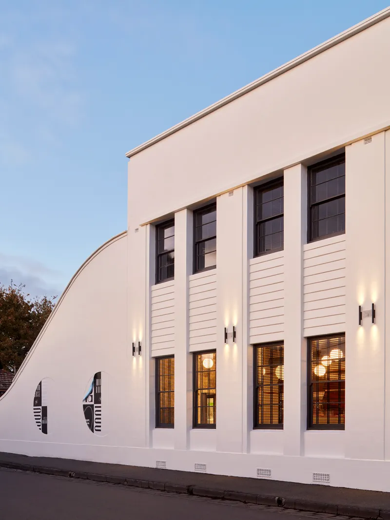 White rendered facade with vertical pilasters and clerestory windows illuminated at dusk from the street