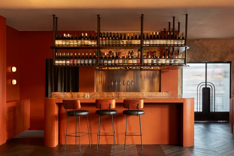 Bar counter with terracotta stone top, suspended shelving, and three bar stools beneath backlit bottles