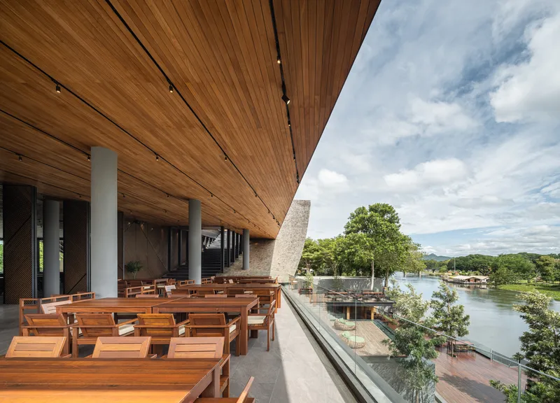 Covered terrace with timber seating and slatted wood ceiling overlooking a river under partly cloudy skies