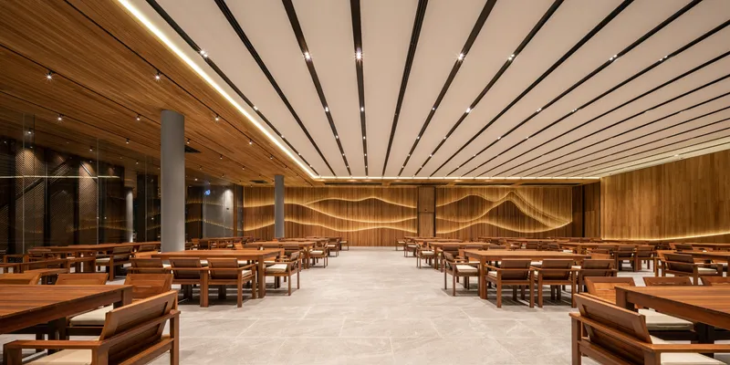 Dining hall with undulating wood relief wall and linear ceiling lighting above timber tables