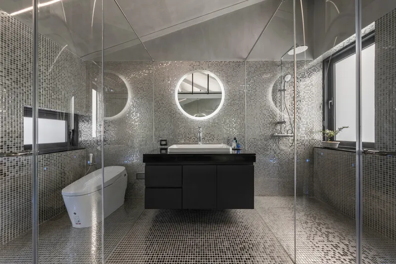 Bathroom clad in reflective silver mosaic tile with glass shower enclosure and round backlit mirrors