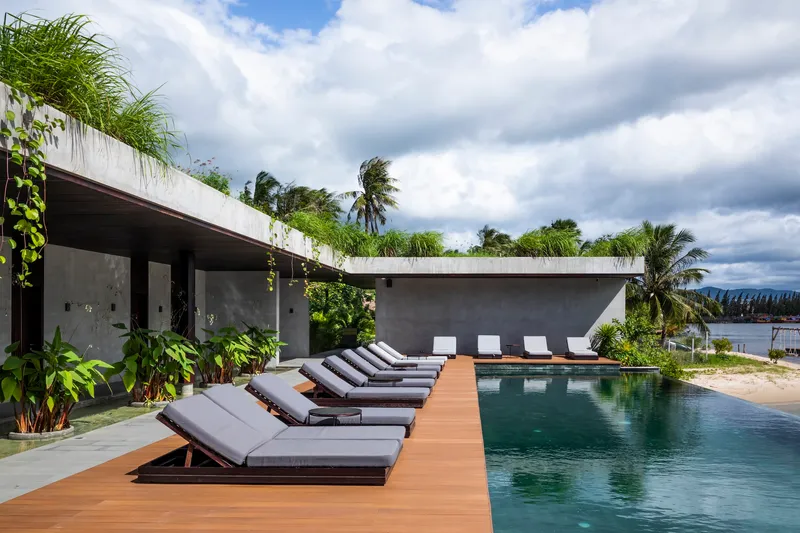 Pool deck with timber planks and loungers beside concrete volumes with green roofs and palms