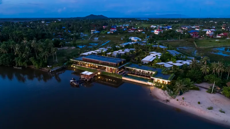 Drone view at dusk showing the illuminated two-story linear buildings alongside the river and sandy beach