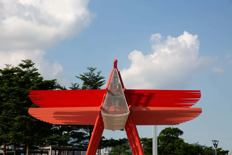 Close-up of the red steel canopy with mesh enclosure and layered horizontal fins against a partly cloudy sky