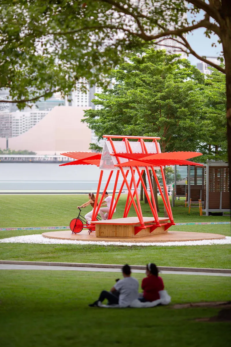 View of the red steel pavilion on a circular white gravel base with two people seated on the lawn