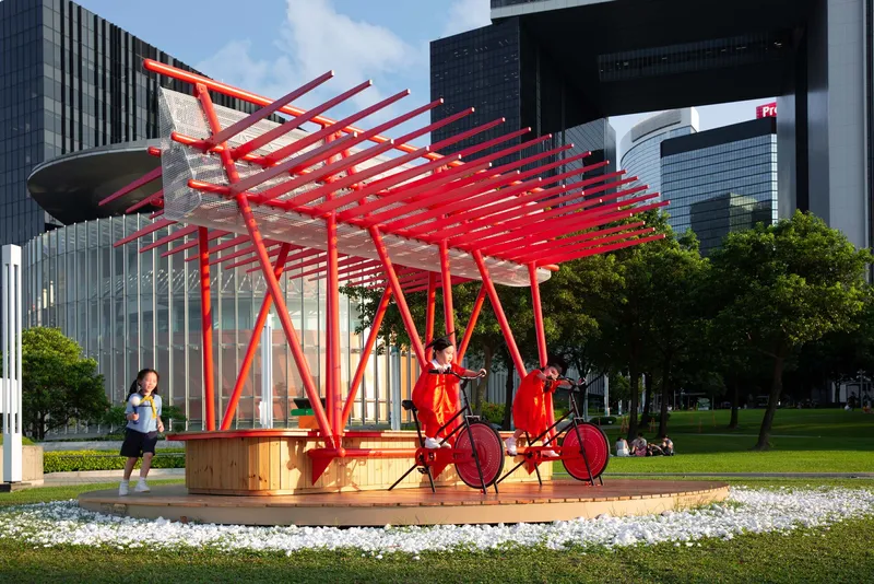The red canopy structure with angled steel supports and bicycles underneath, set against glass towers in afternoon light