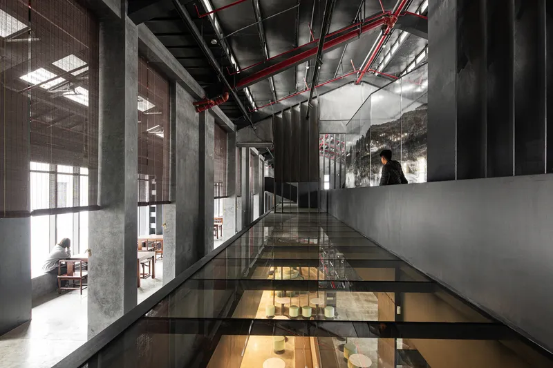 Glass-floored bridge suspended between concrete columns with red conduit overhead and a figure standing at the end