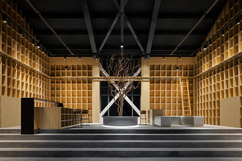 Gallery space with gridded timber wall shelving and a bare tree in a circular planter beneath exposed steel trusses
