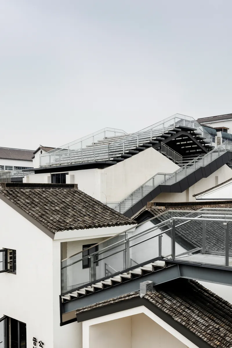 Layered steel staircases zigzagging between white plastered walls and grey-tiled roofs under overcast sky