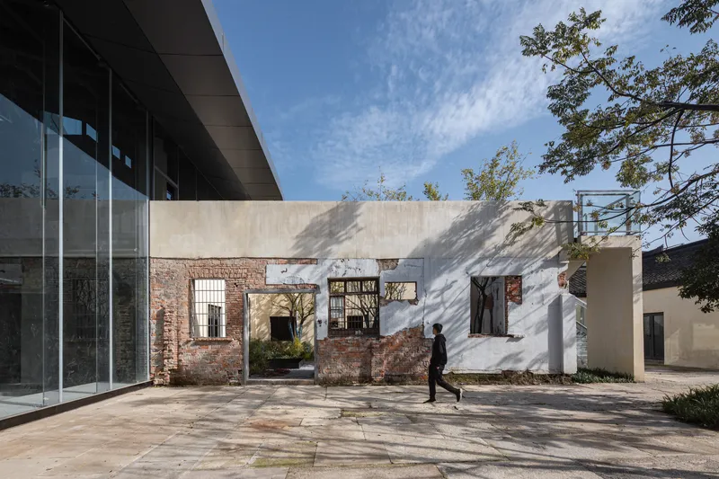 Preserved brick ruin facade with arched openings between white plastered walls as pedestrian walks across paved plaza