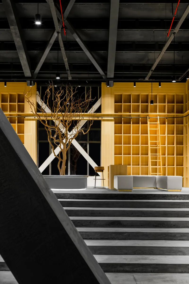 Interior lobby with exposed diagonal steel trusses above yellow illuminated shelving wall and sculptural tree branch