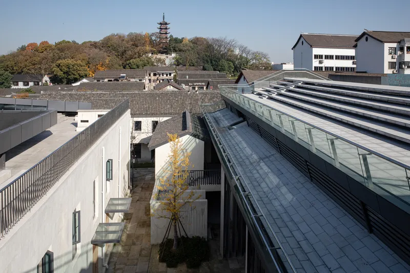 View across sloped glass roofs and courtyard with single tree toward traditional tile roofs