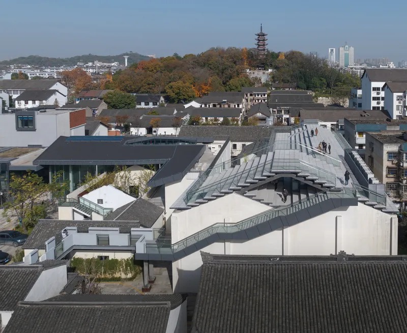 Aerial view of the zigzagging rooftop walkway connecting white volumes among tiled roofs and a distant pagoda