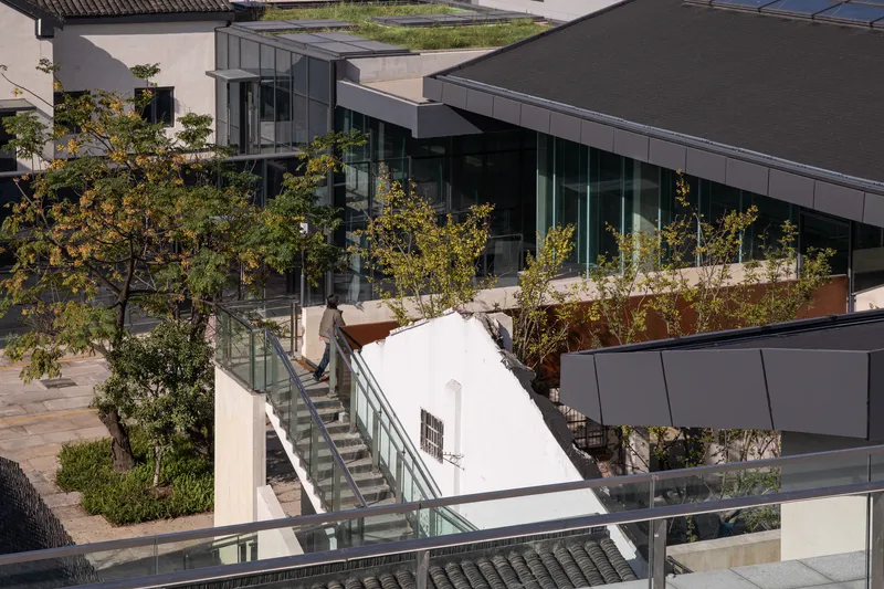 Glass-railed staircase bridging rooftop terraces planted with young trees in afternoon sunlight