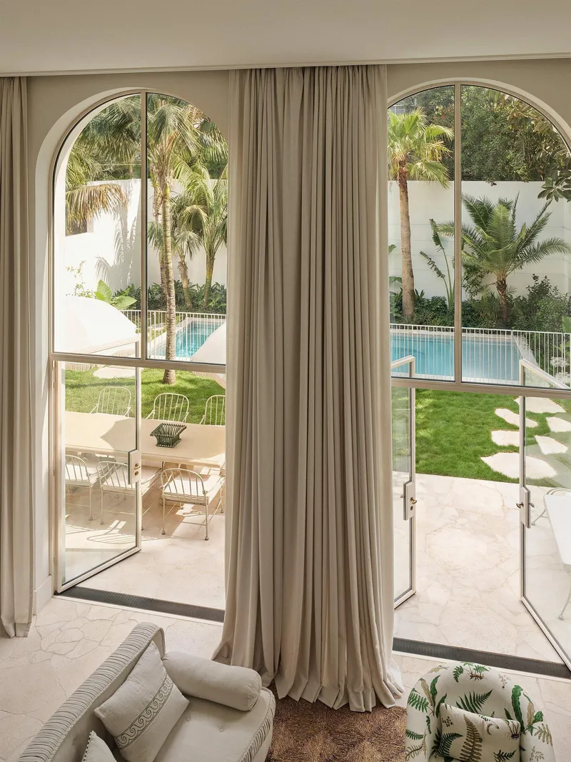 Interior view through arched glass doors with beige curtains overlooking a pool and lawn with palms