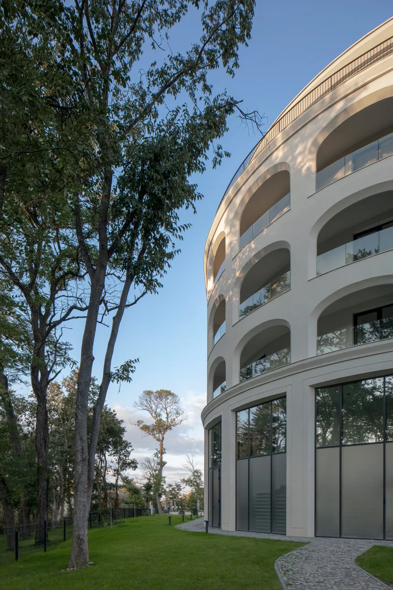 Curved corner of the white facade with stacked arched balconies beside mature trees