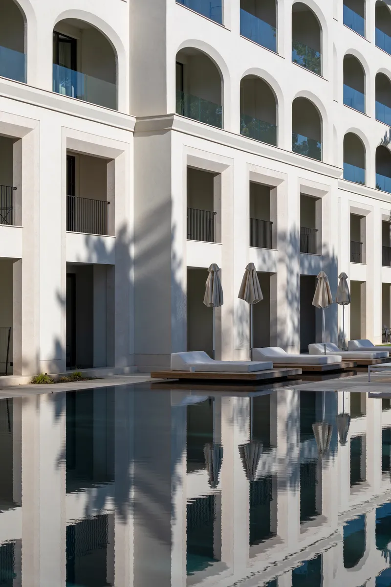 White arched facade reflected in the still pool water with lounge chairs and umbrellas on the deck