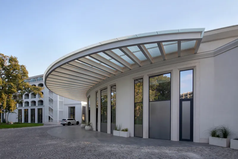 Entry canopy with curved ribbed structure and glazing above tall doors and cobbled forecourt at dusk