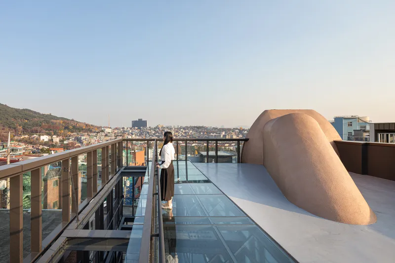 Rooftop terrace with glass floor panels, steel railing, and a person overlooking the hillside neighborhood at dusk