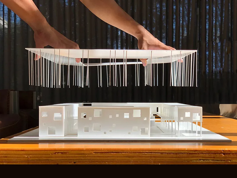 Physical model with hands lifting string-suspended roof structure over white interior volumes below