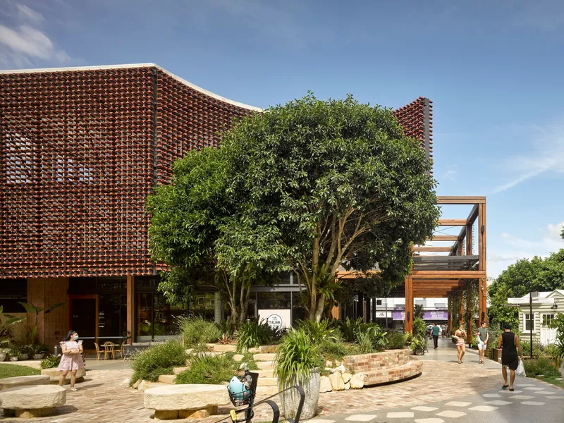 Perforated terracotta brick screen wrapping the facade above a paved plaza with visitors and planted beds