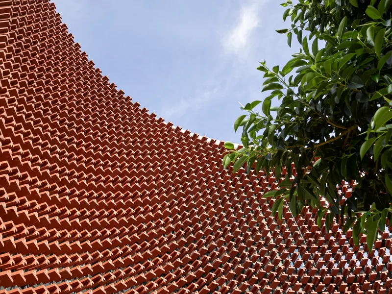 Close-up of the perforated terracotta brick pattern curving against tree foliage and blue sky