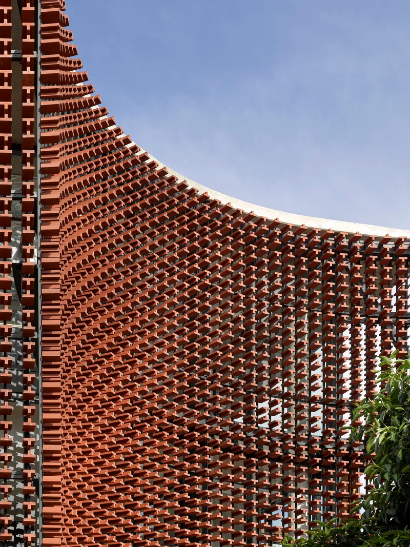 Curving perforated brick screen with staggered coursing against a clear blue sky and vegetation below