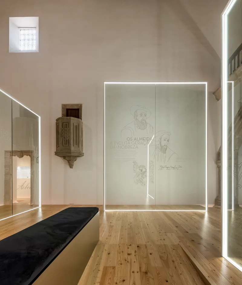 Exhibition room with vaulted ceiling, illuminated glass panels featuring line drawings, and historic stone carved element