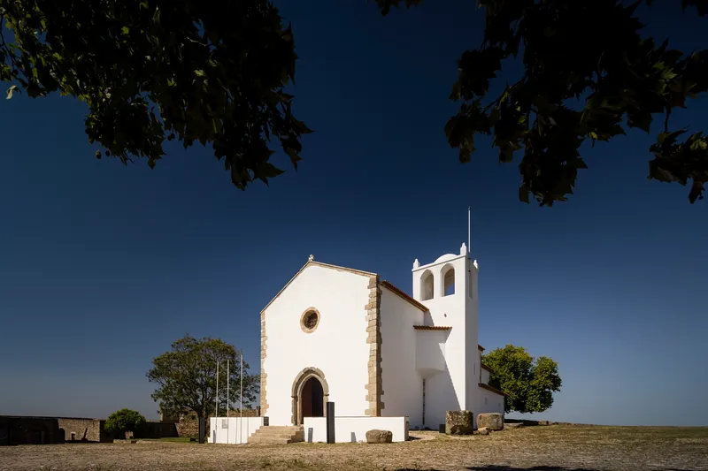 White plastered chapel with bell tower and stone portal on a hilltop under clear blue sky