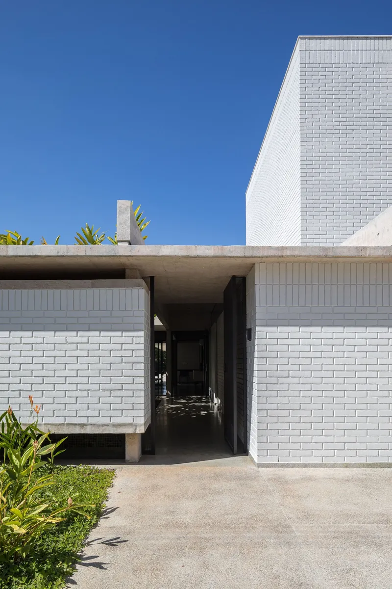 White brick entrance passage beneath an angled concrete soffit with tropical plants beside the path
